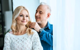 An older couple stands by a window with sheer curtains; the man, in a blue shirt, lovingly touches the woman's blonde hair, while she smiles in a white sweater. The sunlight illuminates their expressions.