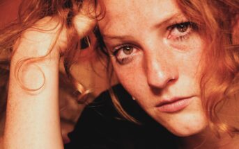 Close-up of a person with curly red hair and freckles holding their head with one hand. Their expression appears serious and intense, with a soft warm light highlighting their features.