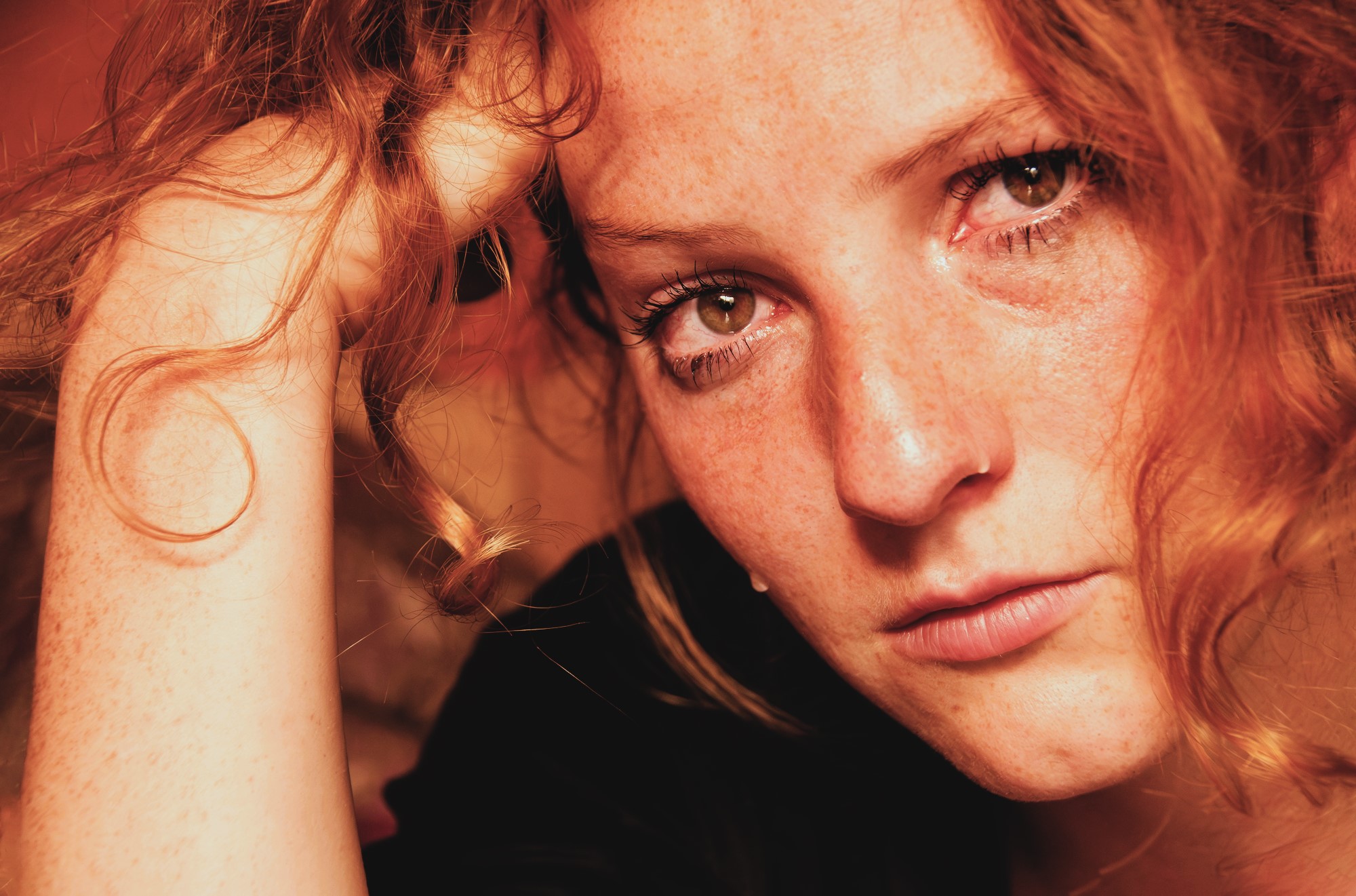 Close-up of a person with curly red hair and freckles holding their head with one hand. Their expression appears serious and intense, with a soft warm light highlighting their features.