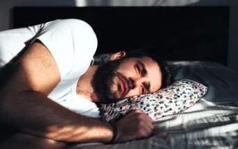 A man with a beard is lying in bed on his side, resting his head on a colorful patterned pillow. He is wearing a white T-shirt and appears thoughtful, as sunlight casts shadows across his face.