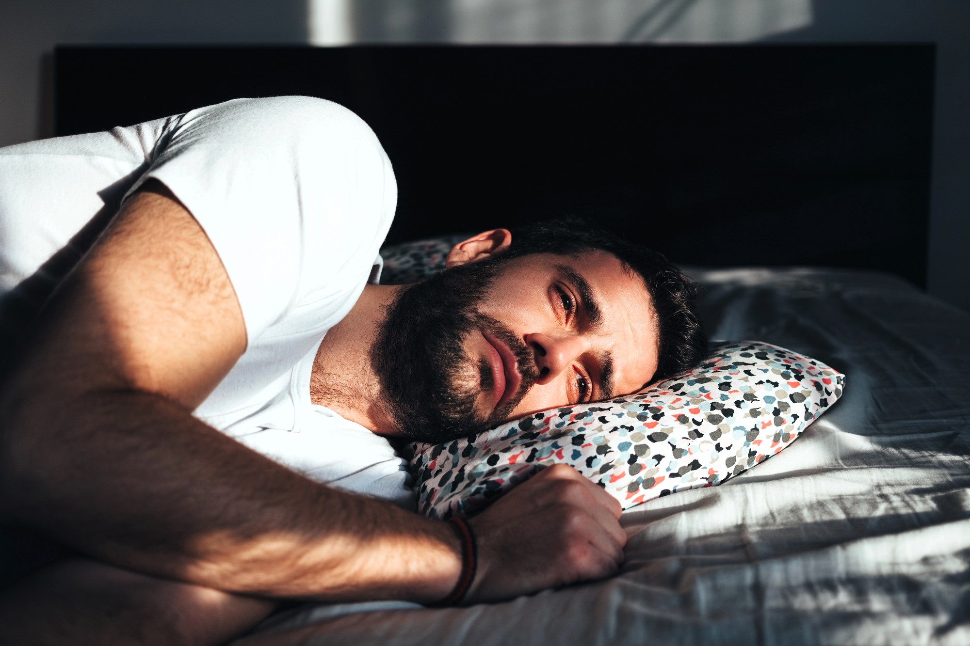 A man with a beard is lying in bed on his side, resting his head on a colorful patterned pillow. He is wearing a white T-shirt and appears thoughtful, as sunlight casts shadows across his face.