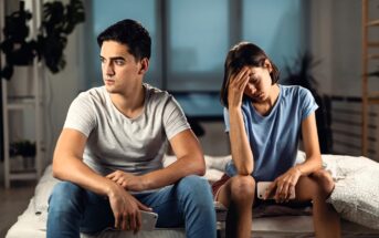 A young man and woman sit on a bed looking upset. The man in a gray shirt and jeans stares to the side, while the woman in a blue shirt holds her head in her hand, appearing distressed. They both hold smartphones.