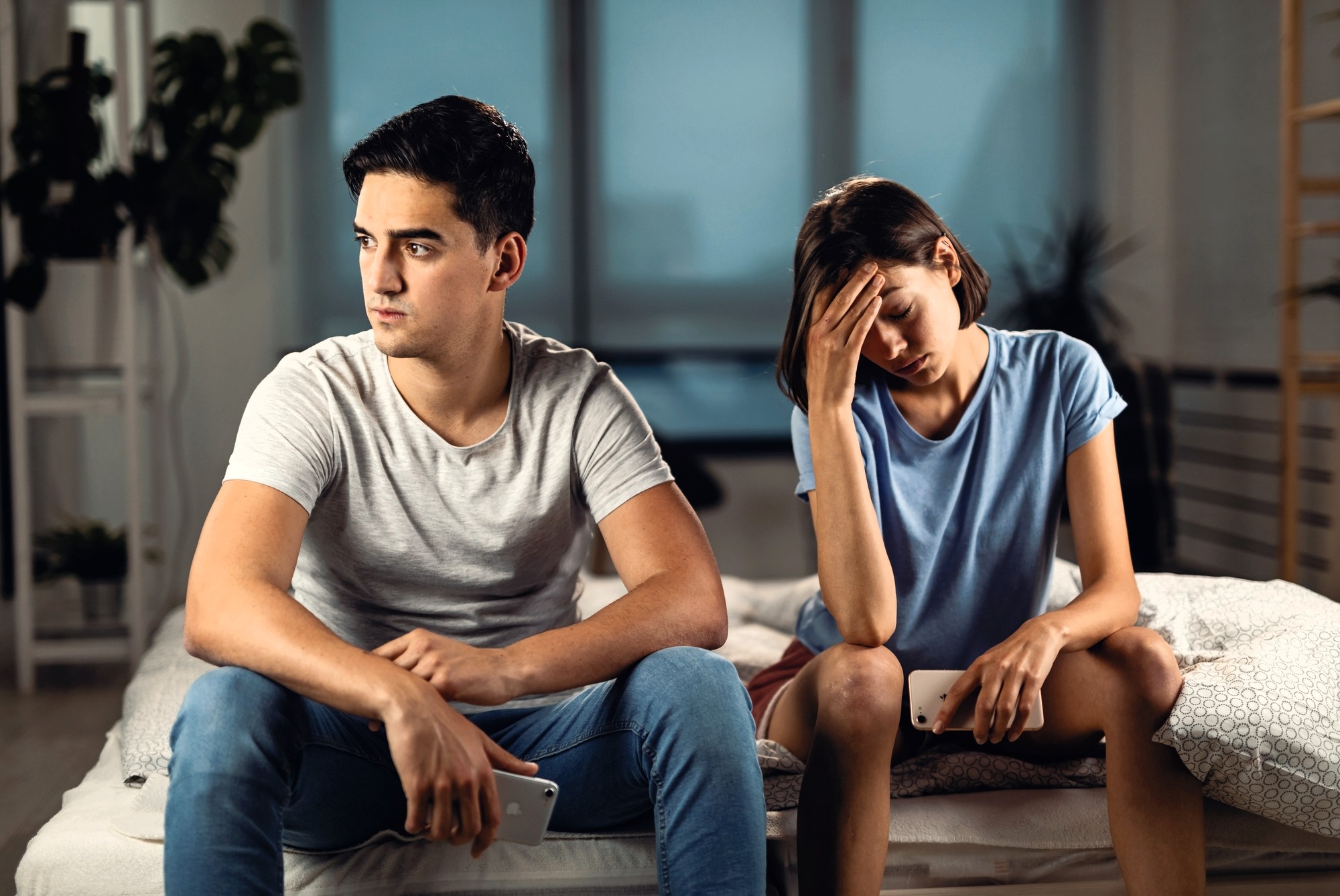 A young man and woman sit on a bed looking upset. The man in a gray shirt and jeans stares to the side, while the woman in a blue shirt holds her head in her hand, appearing distressed. They both hold smartphones.