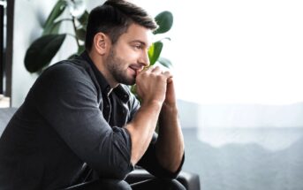 A man with neatly groomed hair and a beard is sitting on a sofa, leaning forward with his hands clasped near his chin. He appears thoughtful and content, looking towards a bright window. Large green plants are visible in the background.