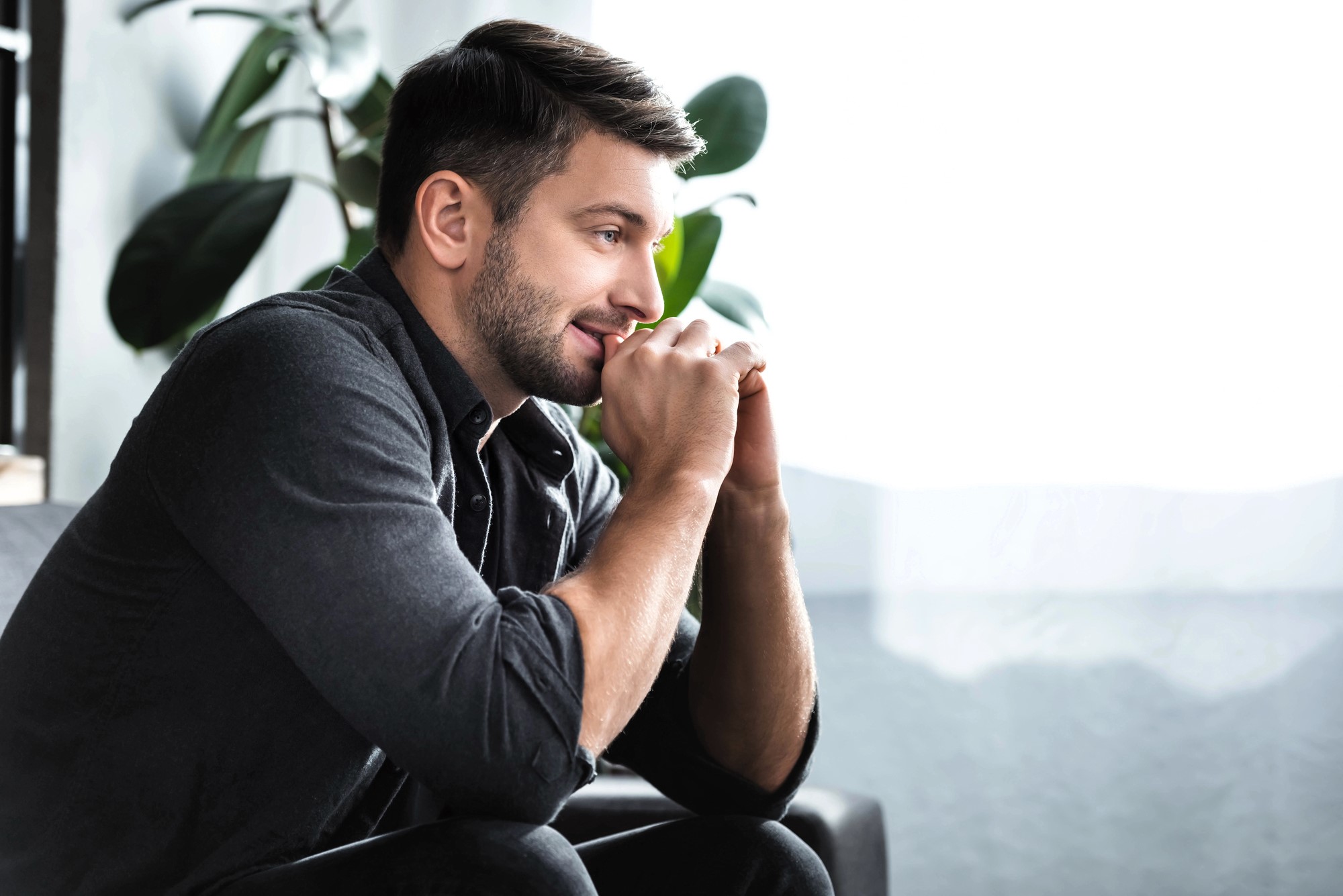 A man with neatly groomed hair and a beard is sitting on a sofa, leaning forward with his hands clasped near his chin. He appears thoughtful and content, looking towards a bright window. Large green plants are visible in the background.