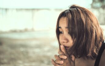 A close-up of a person with chin-length brown hair, looking thoughtful and slightly worried. They have their hands clasped near their face. The background is blurred, featuring neutral tones.