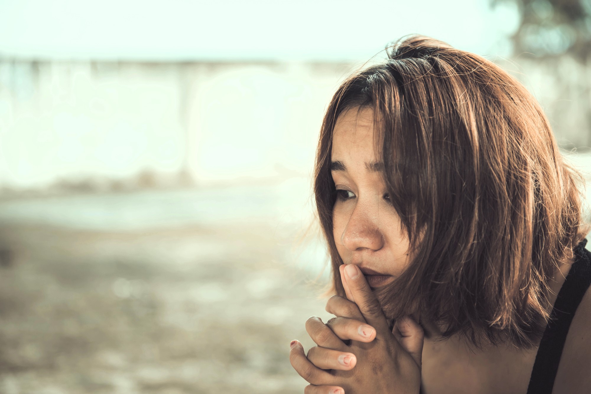 A close-up of a person with chin-length brown hair, looking thoughtful and slightly worried. They have their hands clasped near their face. The background is blurred, featuring neutral tones.