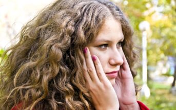 A woman with long, curly hair is holding her face, looking pensive. She is wearing a red top and has pink nail polish. The background is an outdoor setting with green and yellow foliage.