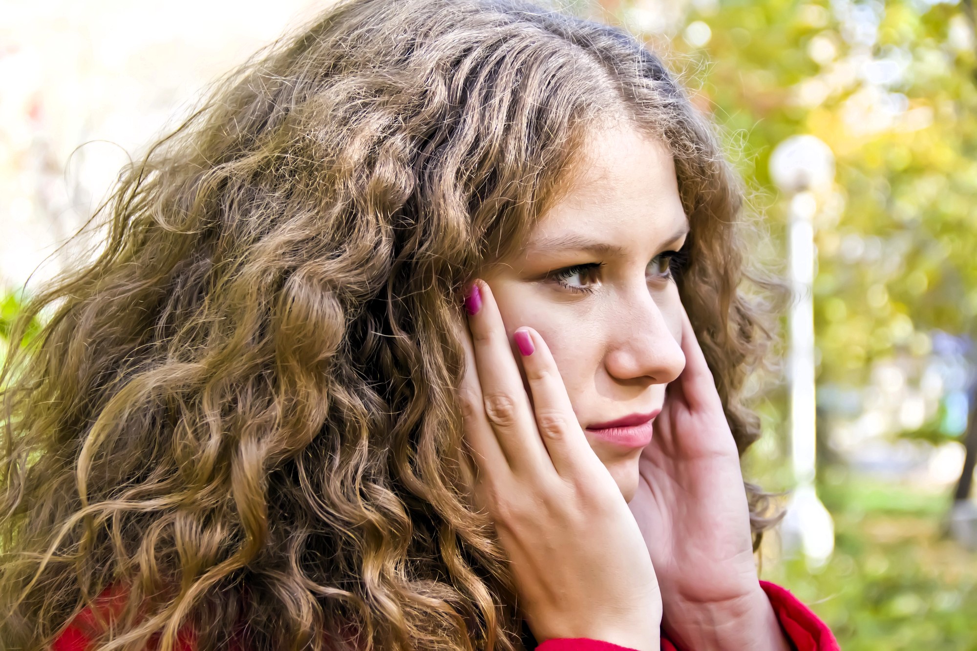 A woman with long, curly hair is holding her face, looking pensive. She is wearing a red top and has pink nail polish. The background is an outdoor setting with green and yellow foliage.