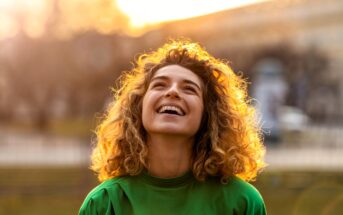 A person with curly hair is smiling and looking upward outdoors. They are wearing a green shirt, and the background is softly blurred with warm sunlight, creating a cheerful and serene atmosphere.