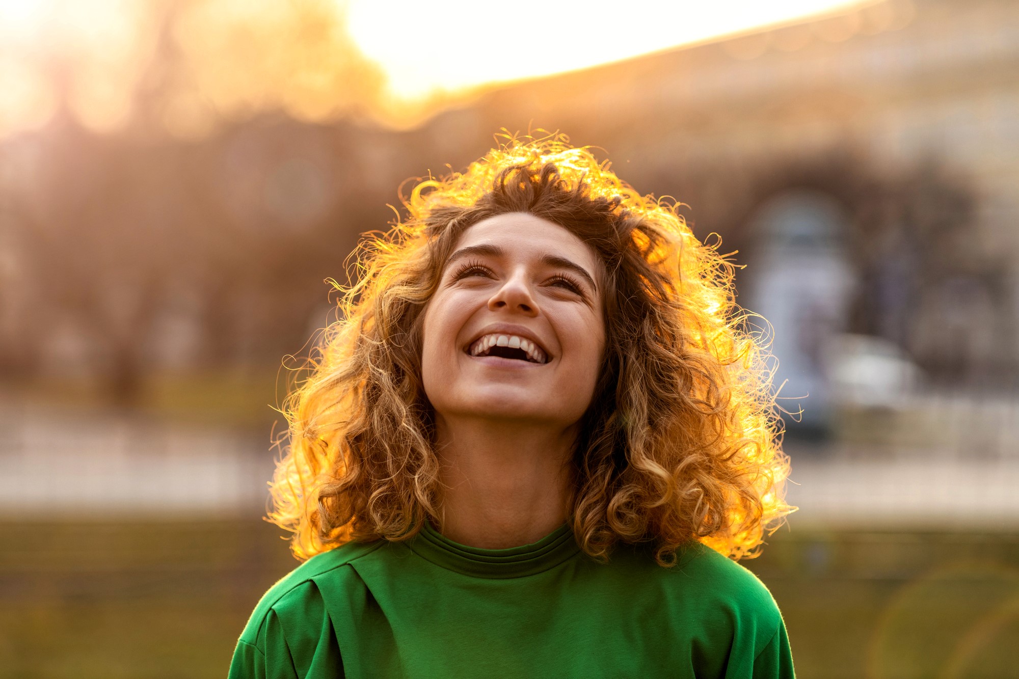 A person with curly hair is smiling and looking upward outdoors. They are wearing a green shirt, and the background is softly blurred with warm sunlight, creating a cheerful and serene atmosphere.