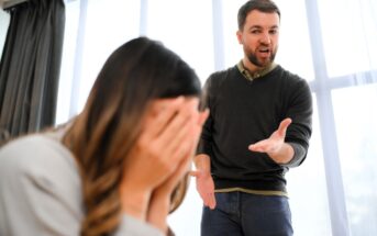 A man with a beard is angrily gesturing towards a woman. The woman is sitting with her face in her hands, appearing upset. They are indoors, with large windows in the background.