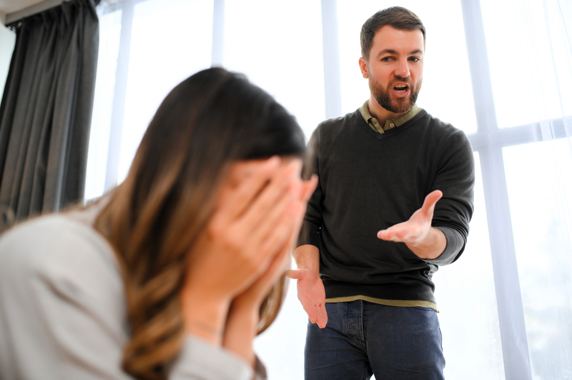 A man with a beard is angrily gesturing towards a woman. The woman is sitting with her face in her hands, appearing upset. They are indoors, with large windows in the background.