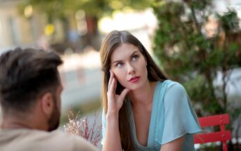 A woman with long brown hair and a blue blouse looks thoughtful while sitting at an outdoor table. She rests her head on her hand. A blurred figure, with short dark hair, sits across from her. The background features greenery.