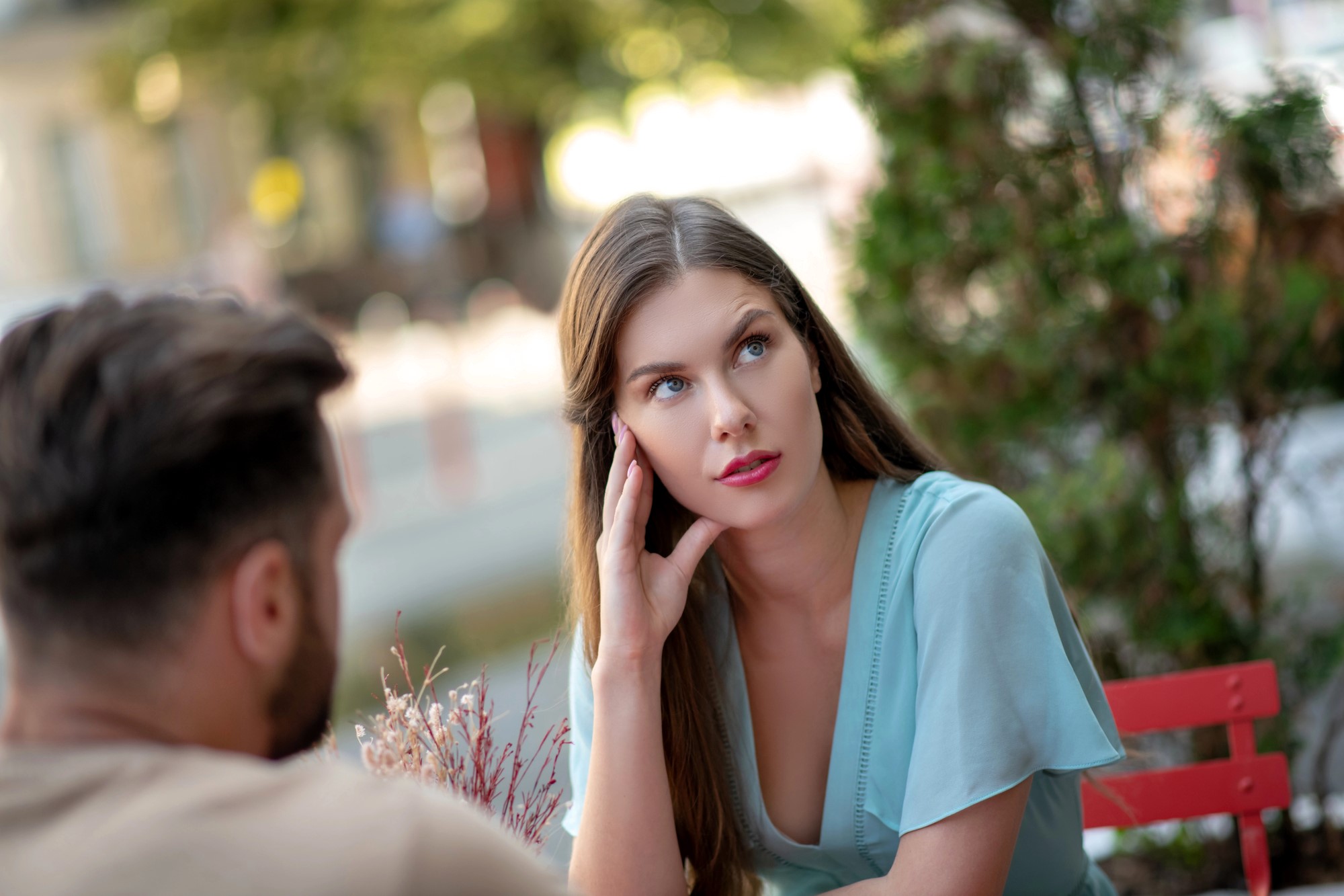 A woman with long brown hair and a blue blouse looks thoughtful while sitting at an outdoor table. She rests her head on her hand. A blurred figure, with short dark hair, sits across from her. The background features greenery.