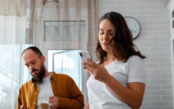 A woman with wavy hair is looking at her smartphone while a man with a beard, wearing a brown shirt, holds a cup and looks at his phone. They are standing in a bright room with white brick walls and a wall clock.