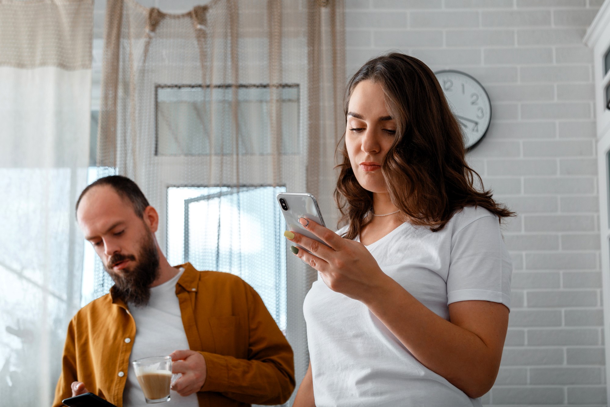 A woman with wavy hair is looking at her smartphone while a man with a beard, wearing a brown shirt, holds a cup and looks at his phone. They are standing in a bright room with white brick walls and a wall clock.