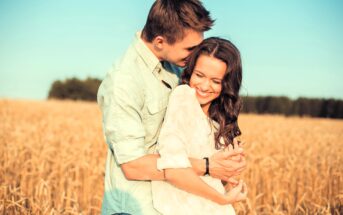 A couple stands in a golden wheat field under a clear blue sky. The man, wearing a light green shirt, embraces the woman from behind. She wears a white blouse and smiles, leaning into him. Trees line the horizon in the background.