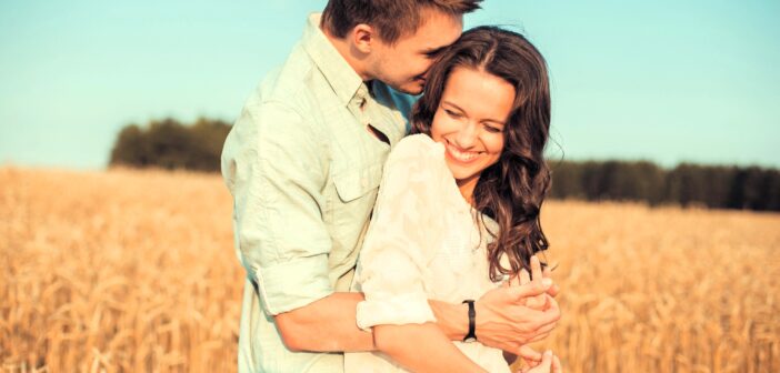 A couple stands in a golden wheat field under a clear blue sky. The man, wearing a light green shirt, embraces the woman from behind. She wears a white blouse and smiles, leaning into him. Trees line the horizon in the background.