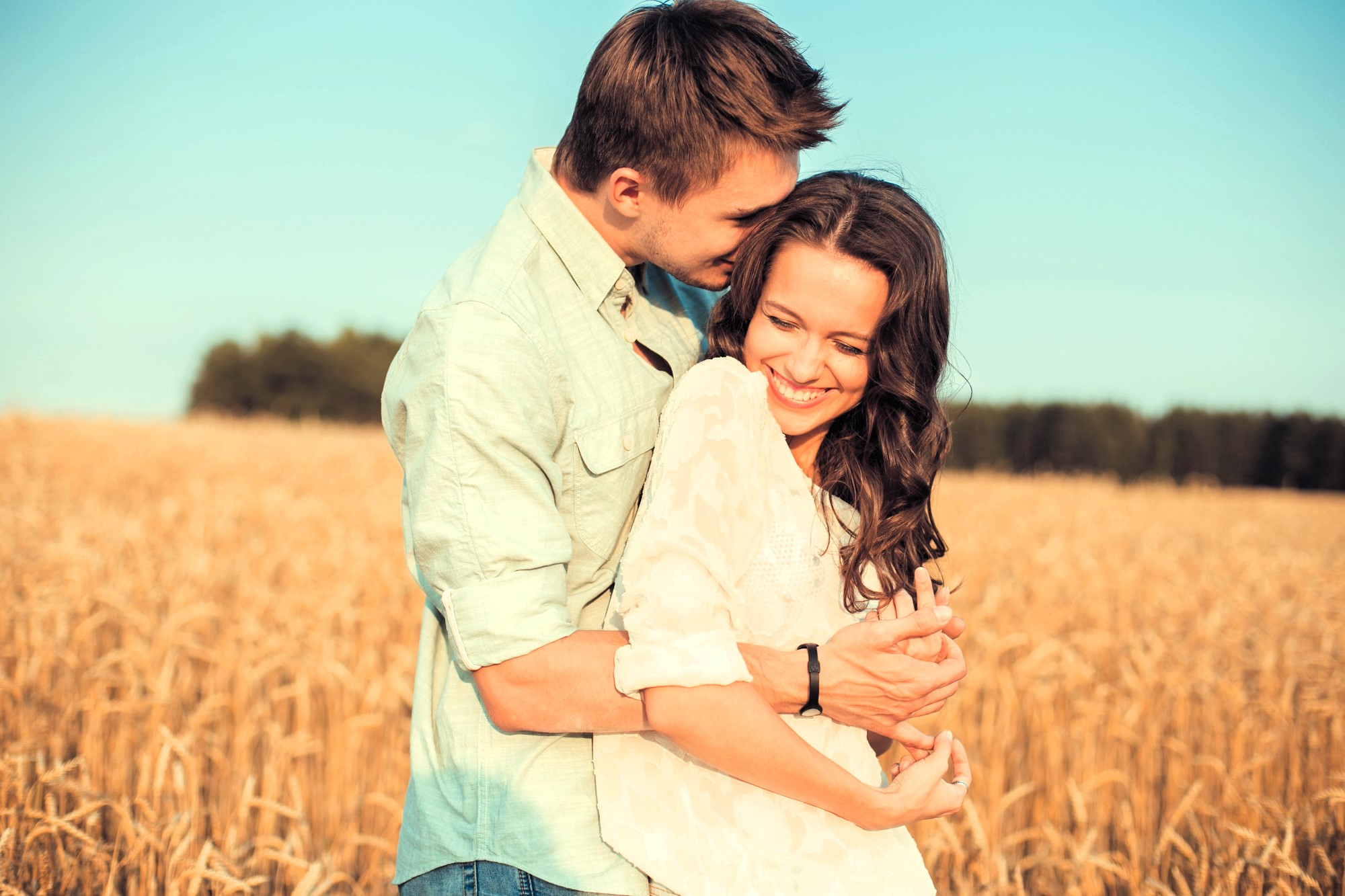 A couple stands in a golden wheat field under a clear blue sky. The man, wearing a light green shirt, embraces the woman from behind. She wears a white blouse and smiles, leaning into him. Trees line the horizon in the background.