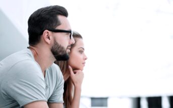 Man and woman sitting side by side, looking intently into the distance. The man has a beard and is wearing glasses and a light-colored shirt. The woman rests her chin on her hands. The background is bright and blurred.