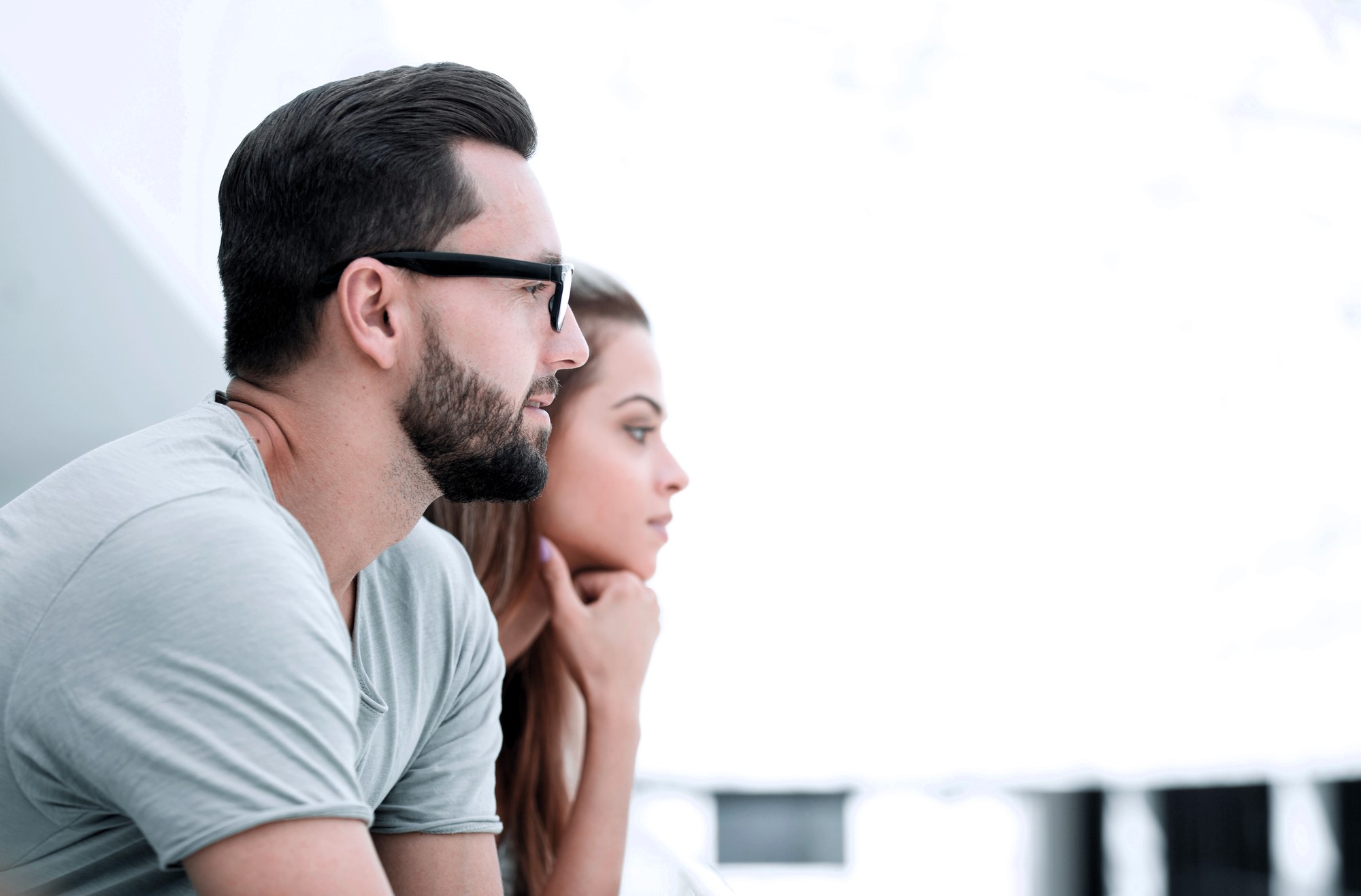 Man and woman sitting side by side, looking intently into the distance. The man has a beard and is wearing glasses and a light-colored shirt. The woman rests her chin on her hands. The background is bright and blurred.