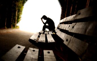 Silhouette of a person sitting on a bench in a tunnel, head resting on hand in a contemplative pose. The entrance is bright, creating a contrast with the shadowy tunnel. The focus is on the person, conveying a sense of solitude.