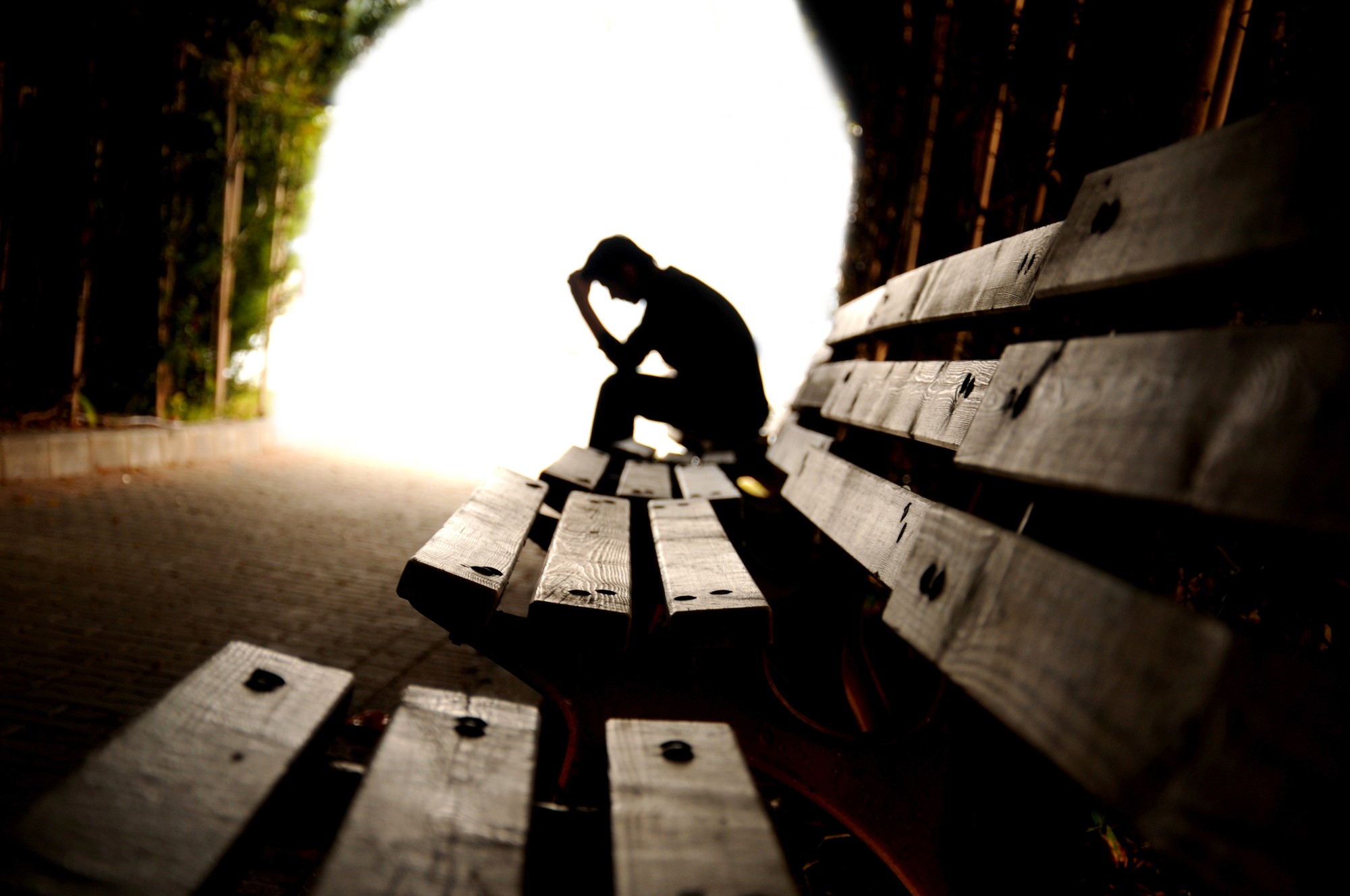 Silhouette of a person sitting on a bench in a tunnel, head resting on hand in a contemplative pose. The entrance is bright, creating a contrast with the shadowy tunnel. The focus is on the person, conveying a sense of solitude.