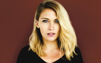 A person with long blonde hair wearing a black top looks directly at the camera with a neutral expression. The background is a solid maroon color.