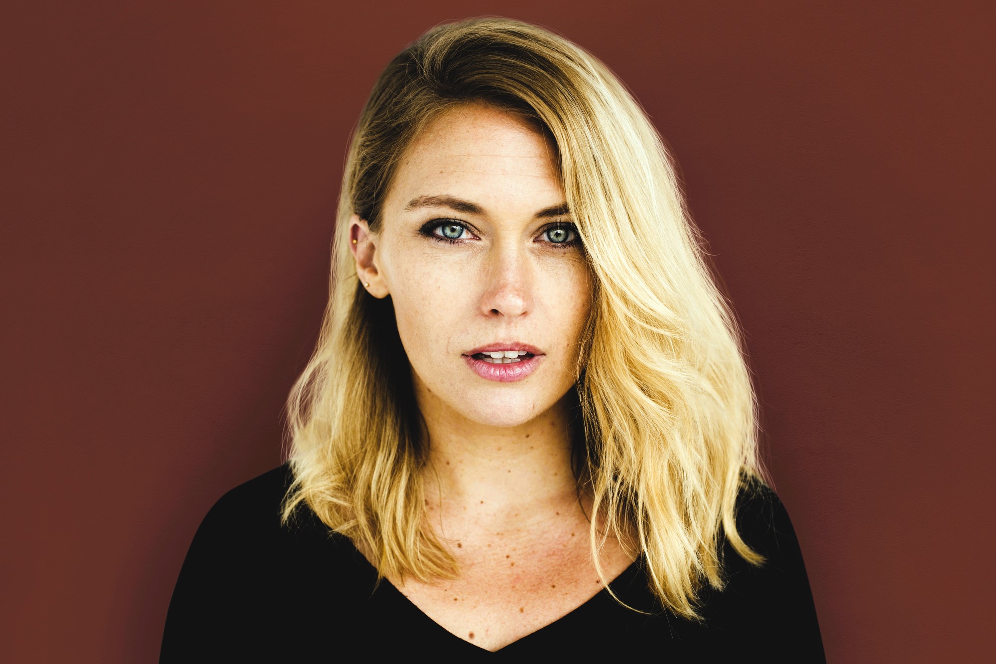 A person with long blonde hair wearing a black top looks directly at the camera with a neutral expression. The background is a solid maroon color.