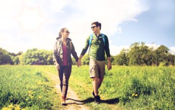 A couple walks hand in hand on a sunny day through a grassy field. Both wear sunglasses and casual outdoor clothing. The sky is clear with a few clouds and a bright sun. Trees line the background.