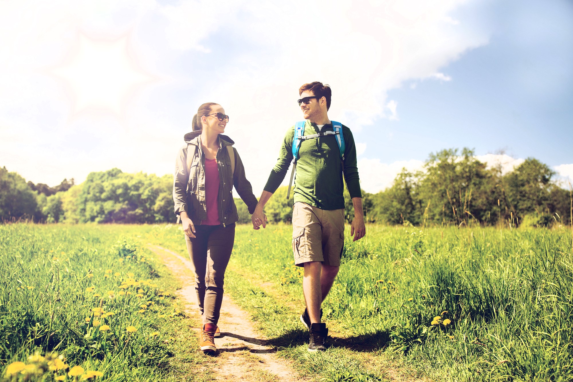 A couple walks hand in hand on a sunny day through a grassy field. Both wear sunglasses and casual outdoor clothing. The sky is clear with a few clouds and a bright sun. Trees line the background.