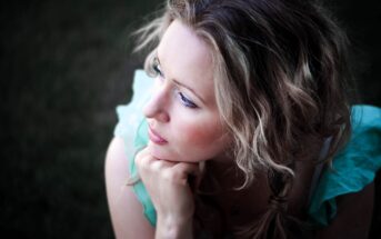 A woman with wavy blonde hair looks thoughtfully into the distance. She rests her chin on her hand, wearing a light blue, ruffled top. The background is dark and blurred, creating a serene and contemplative atmosphere.