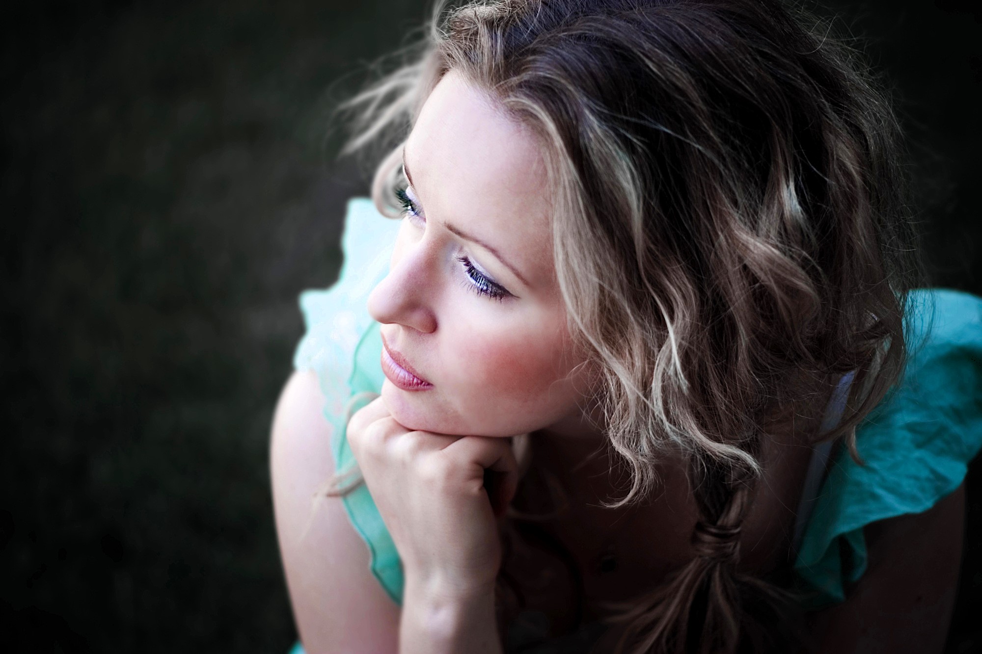 A woman with wavy blonde hair looks thoughtfully into the distance. She rests her chin on her hand, wearing a light blue, ruffled top. The background is dark and blurred, creating a serene and contemplative atmosphere.