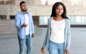 A woman in a white shirt and light blue cardigan walks away confidently with a neutral expression, while a man in a blue shirt and jeans stands behind her, gesturing with one hand. They are outdoors in an urban setting.