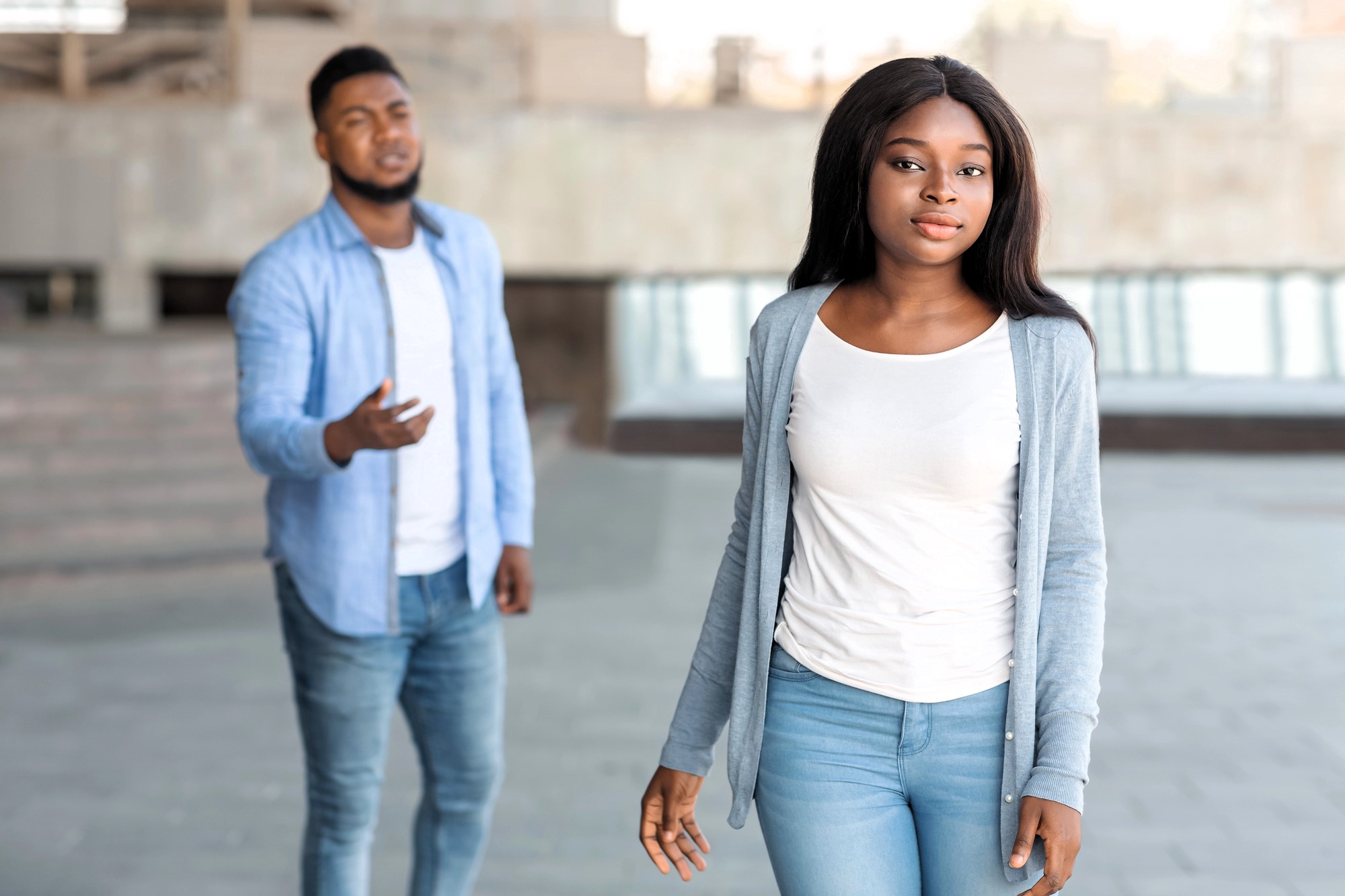 A woman in a white shirt and light blue cardigan walks away confidently with a neutral expression, while a man in a blue shirt and jeans stands behind her, gesturing with one hand. They are outdoors in an urban setting.