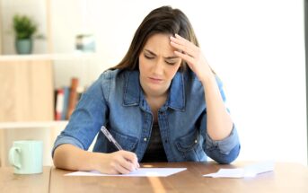 A woman sits at a table, looking stressed while writing on a piece of paper. Her hand supports her head, and a mug is placed on the table. A bookshelf with plants and books is in the background.