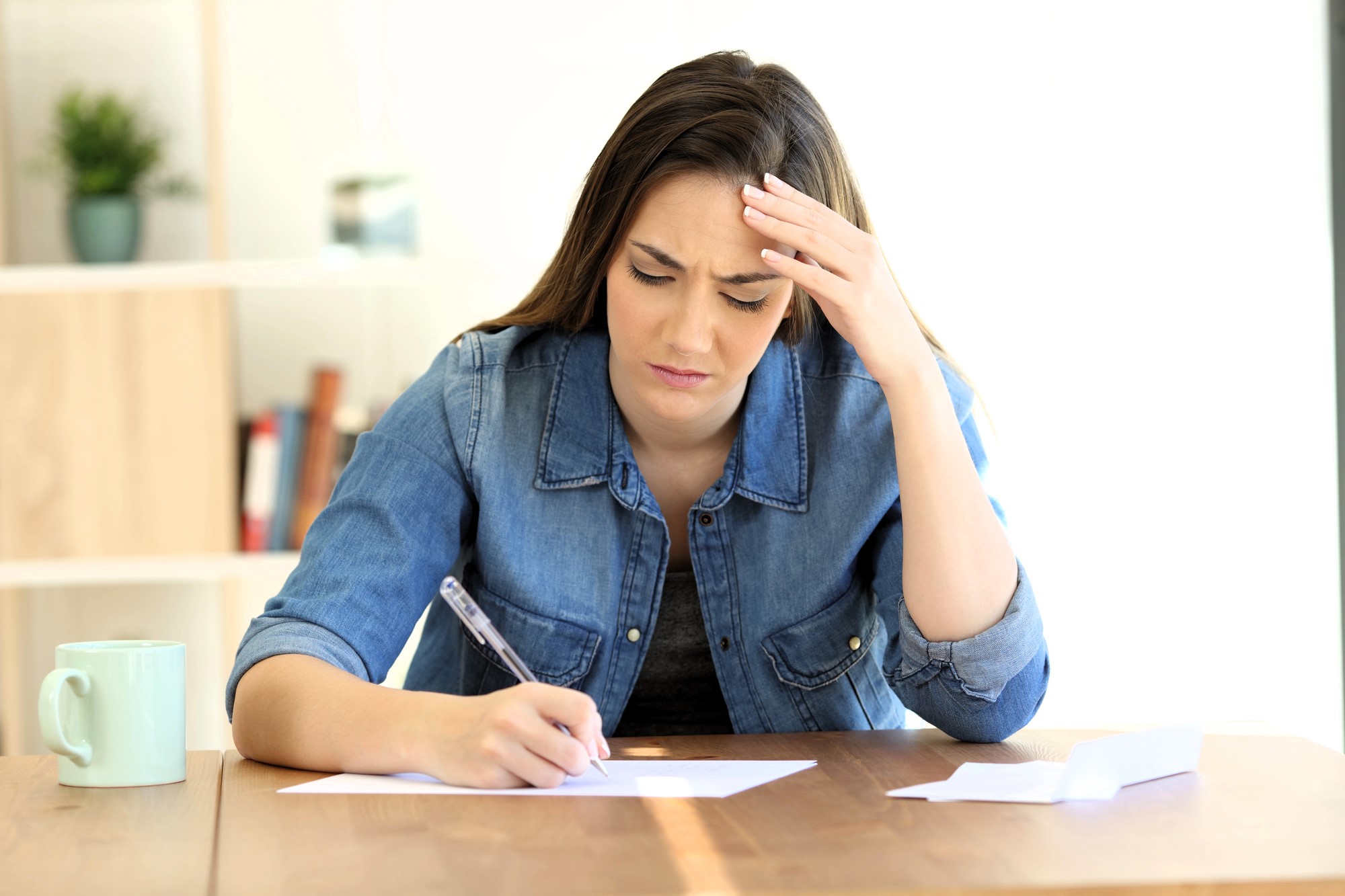 A woman sits at a table, looking stressed while writing on a piece of paper. Her hand supports her head, and a mug is placed on the table. A bookshelf with plants and books is in the background.