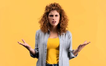 A woman with curly hair looks confused, standing against a yellow background. She is wearing a gray shirt over a yellow top and jeans, with her hands raised slightly to the sides, palms up.