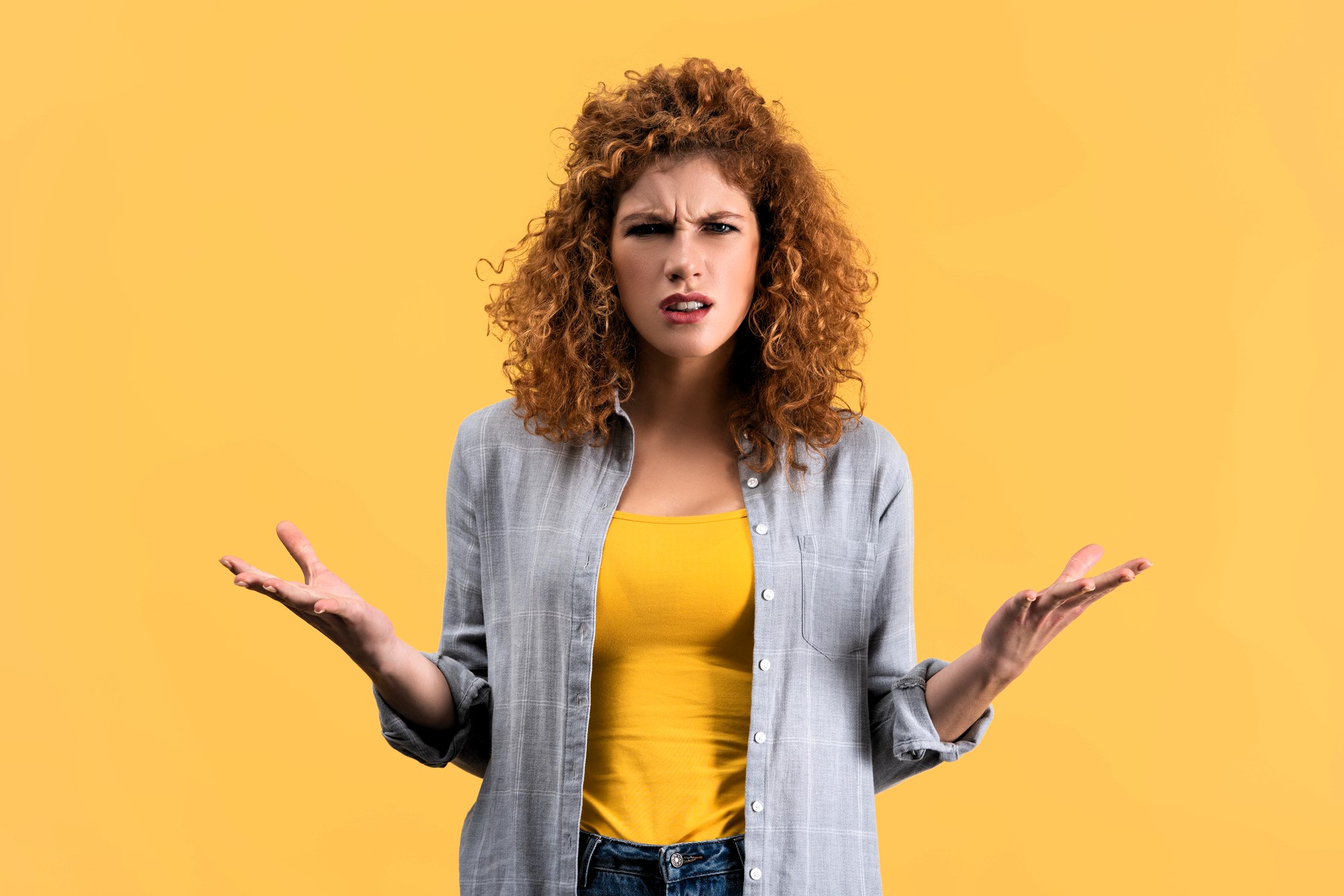 A woman with curly hair looks confused, standing against a yellow background. She is wearing a gray shirt over a yellow top and jeans, with her hands raised slightly to the sides, palms up.