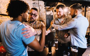 Four men in casual clothing are in a bar, shaking hands and smiling. They are surrounded by tall glasses of beer on a table in a warmly lit, rustic setting. The mood is friendly and upbeat.