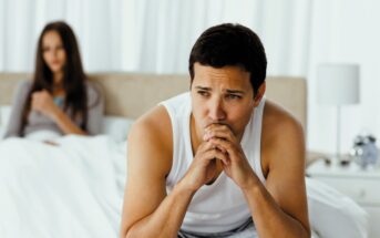 A man sits on the edge of a bed looking thoughtful and concerned, resting his chin on his hands. In the background, a woman sits on the bed, partially blurred, with her hands folded across her chest. The room is softly lit.