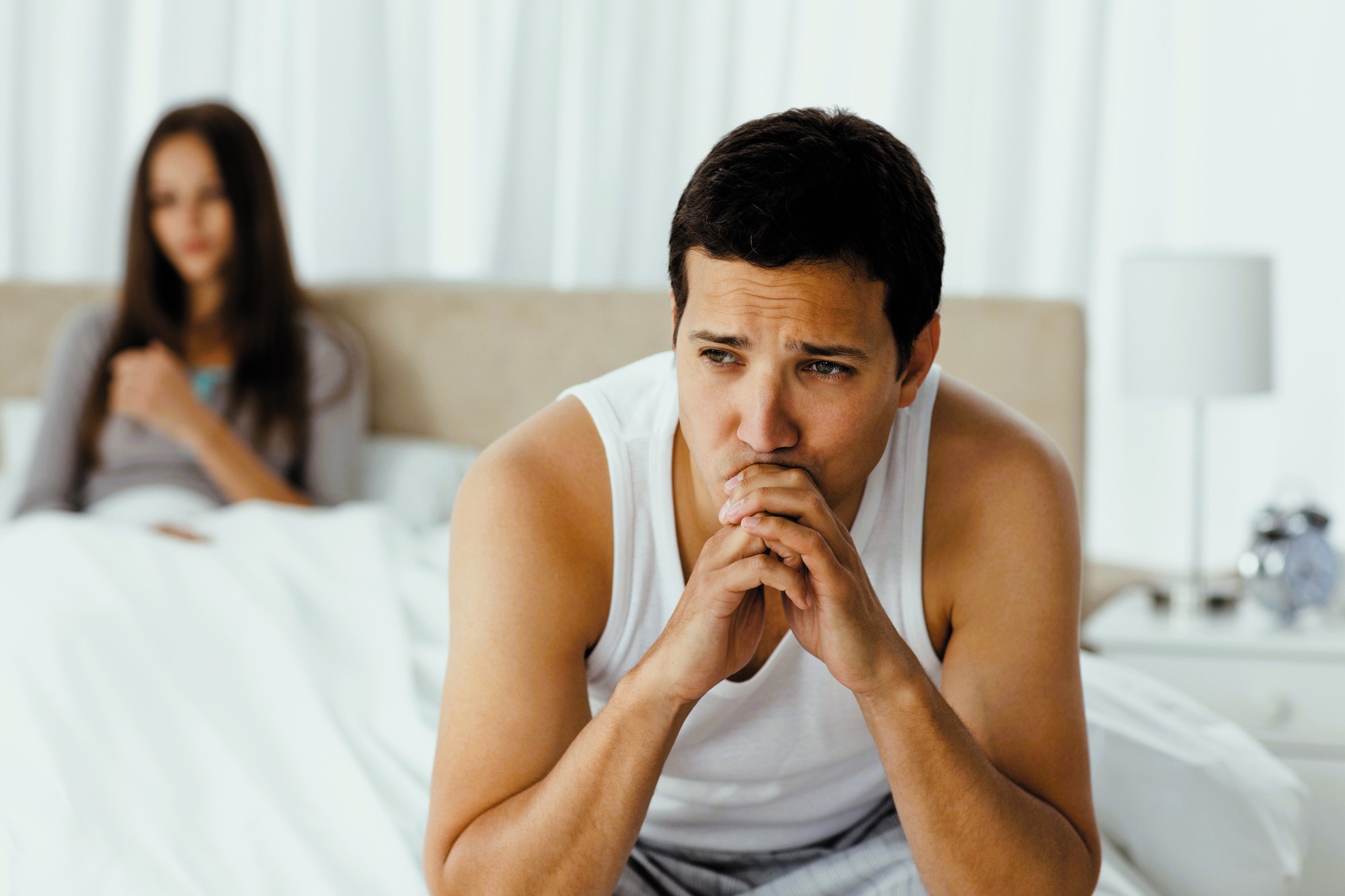 A man sits on the edge of a bed looking thoughtful and concerned, resting his chin on his hands. In the background, a woman sits on the bed, partially blurred, with her hands folded across her chest. The room is softly lit.