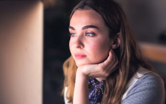 A woman with long brown hair and a thoughtful expression rests her chin on her hand while looking into the distance. She's wearing a gray top and a blue scarf with yellow dots. The background is softly blurred.