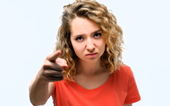 A woman with curly blonde hair wearing an orange shirt points a finger forward with an intense expression against a plain background.