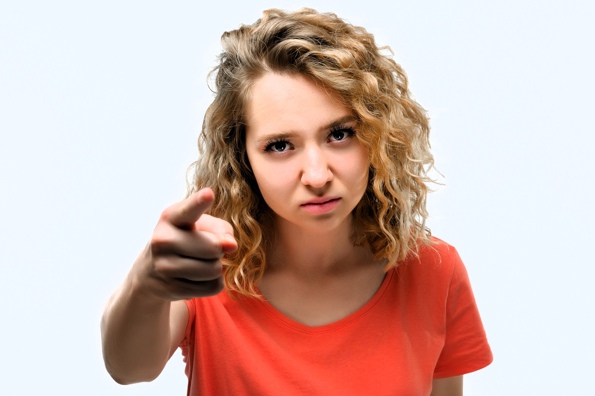 A woman with curly blonde hair wearing an orange shirt points a finger forward with an intense expression against a plain background.