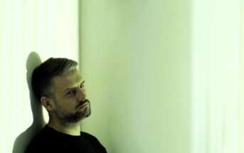 A man with short hair and a beard is leaning against a white wall, gazing off to the side with a thoughtful expression. The lighting casts a greenish hue across the scene, creating a moody atmosphere.