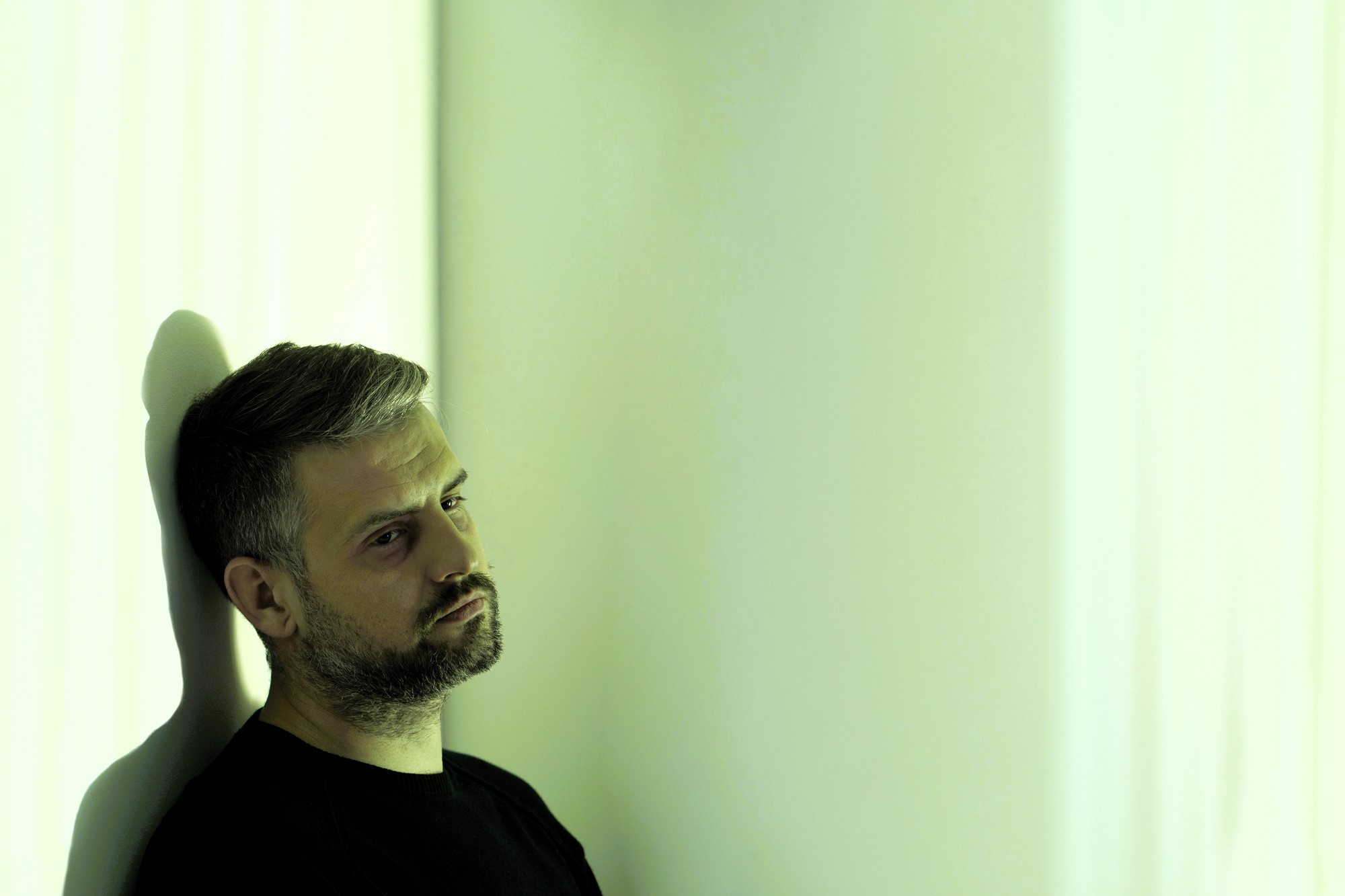 A man with short hair and a beard is leaning against a white wall, gazing off to the side with a thoughtful expression. The lighting casts a greenish hue across the scene, creating a moody atmosphere.