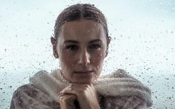 Person with a thoughtful expression stands behind a rain-streaked window, wearing a cozy sweater. They rest their chin on their hands, gazing outward. The blurred background gives a moody, reflective atmosphere.