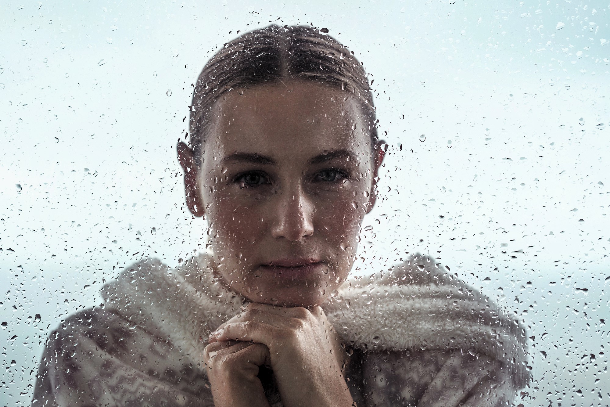 Person with a thoughtful expression stands behind a rain-streaked window, wearing a cozy sweater. They rest their chin on their hands, gazing outward. The blurred background gives a moody, reflective atmosphere.